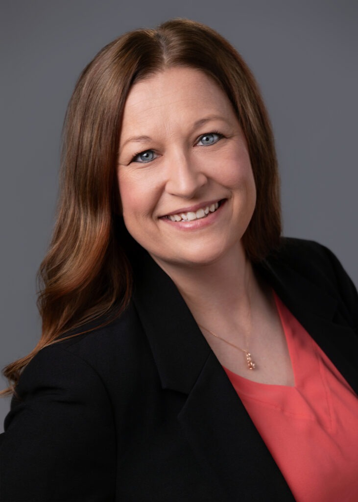 Smiling woman with long brown hair and blue eyes, wearing a black blazer over a coral blouse in a gray studio backdrop.