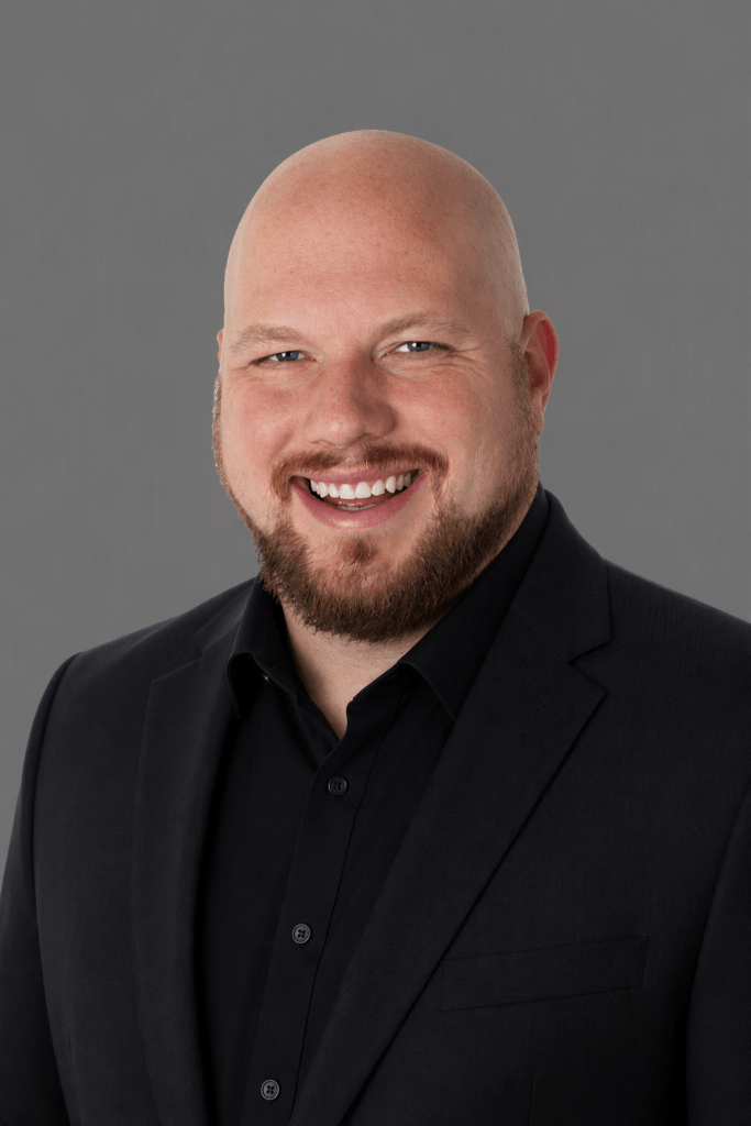 Smiling bald man in a black blazer and shirt against a gray backdrop (professional headshot).