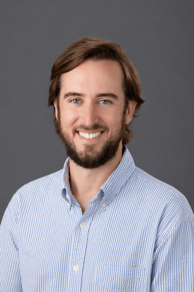 Smiling man with brown hair and beard in a blue striped button-down shirt against a gray backdrop.