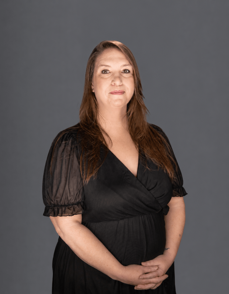 Portrait of a smiling woman with long brown hair in a black wrap dress against a gray studio backdrop.