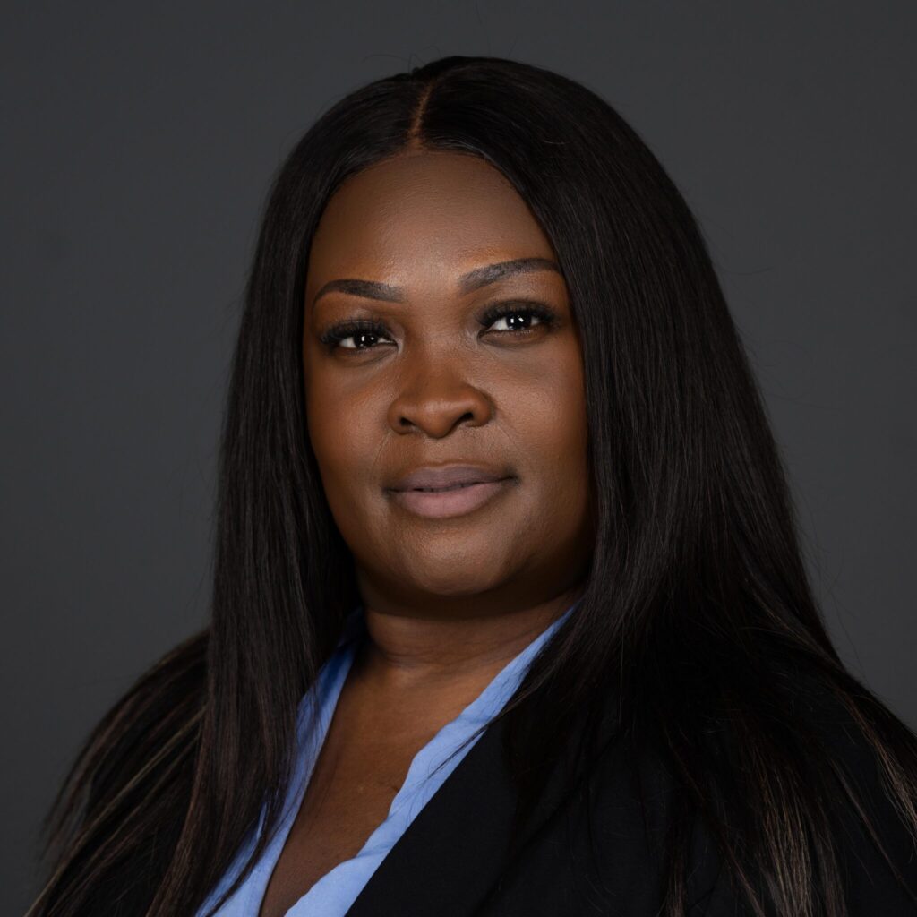 Close-up portrait of a Black woman with long straight hair, wearing a blue blouse and black blazer against a dark gray background.
