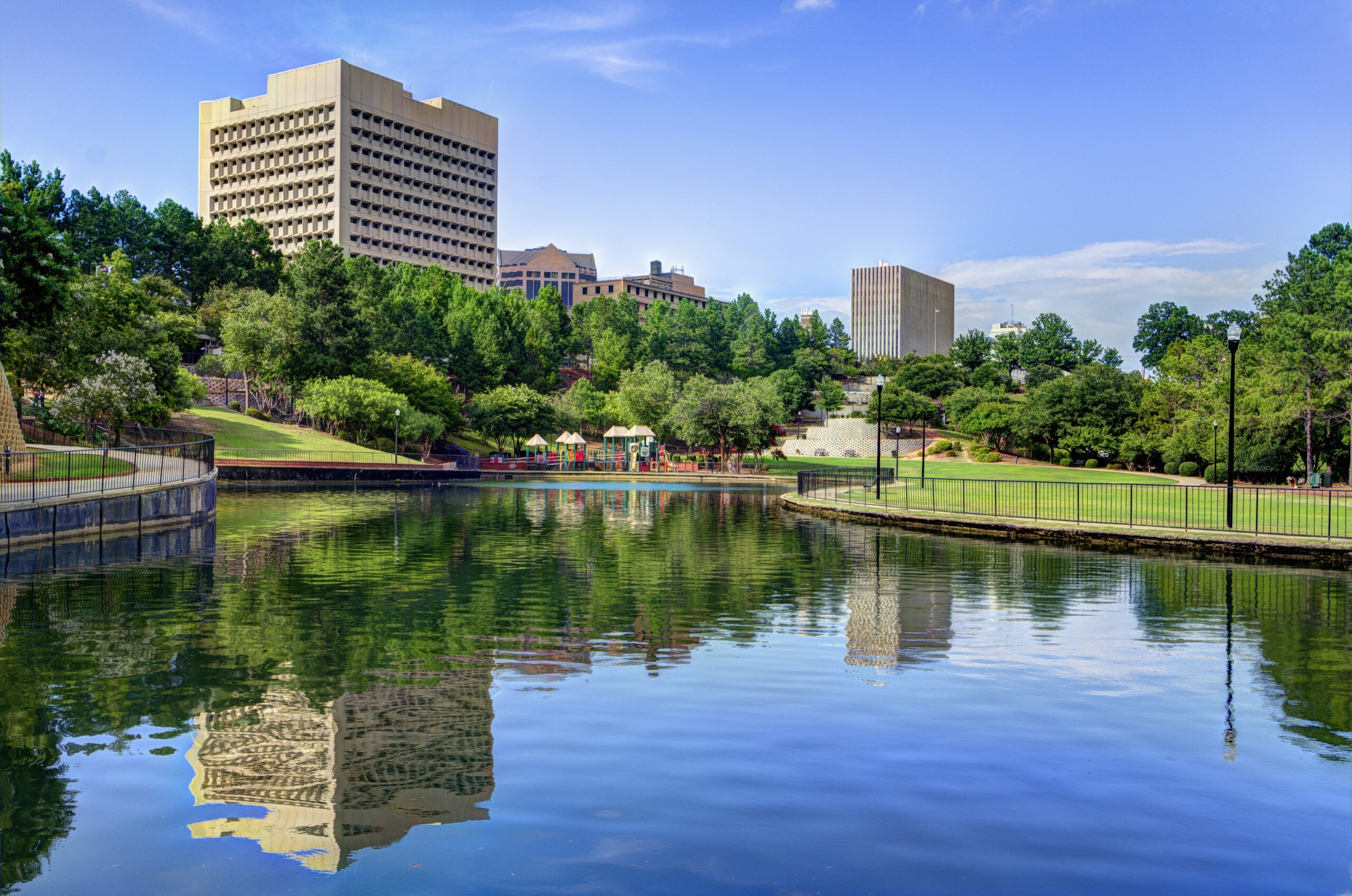 City skyline with tall beige office buildings behind a green park, reflected in calm water.