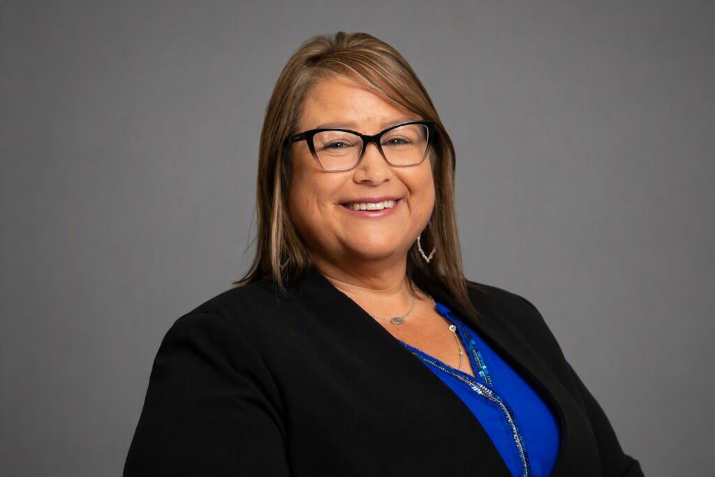 Smiling woman with glasses in a black blazer and blue blouse against a gray backdrop (professional headshot).