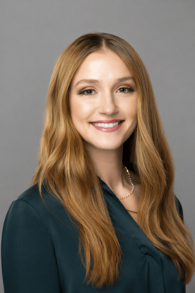 Portrait of a smiling woman with long wavy light brown hair, wearing a teal blouse and pearl necklace, against a gray backdrop.
