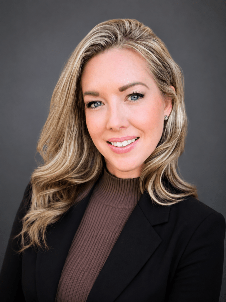 Professional headshot of a smiling blonde woman in a brown turtleneck and black blazer against a gray backdrop.