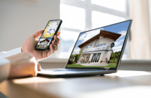 A person holds a smartphone showing a property map next to a laptop displaying a two‑story house in a bright room.