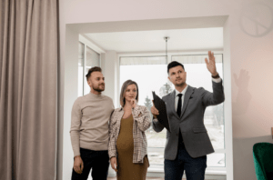 Real estate agent in a gray suit explains features to a couple inside a bright room with a large window, clipboard in hand, gesturing toward something they’re viewing.