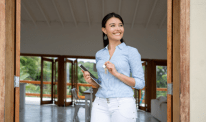 Smiling woman standing in a doorway, holding a tablet and keys, wearing a light blue striped shirt and white jeans.